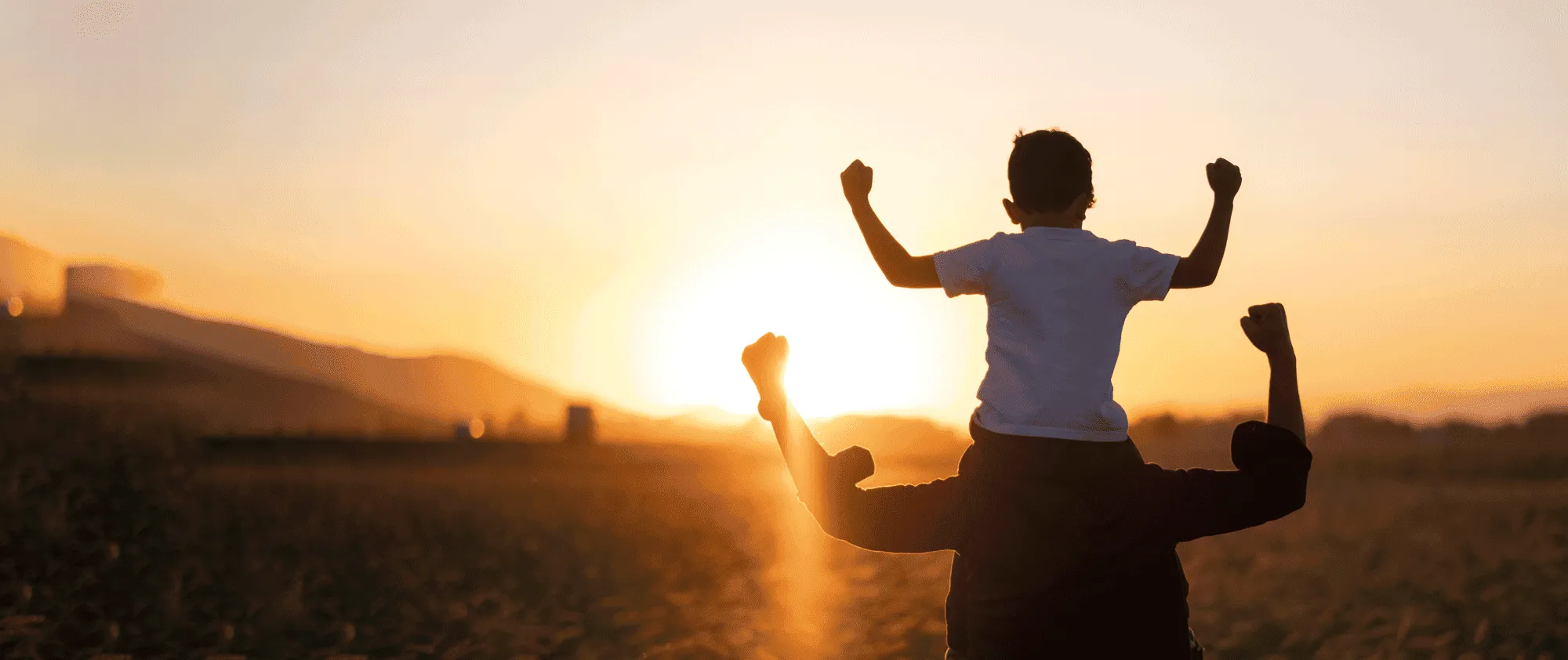 Kind auf Schultern eines Erwachsenen hebt die Fäuste triumphierend bei Sonnenuntergang am Strand.