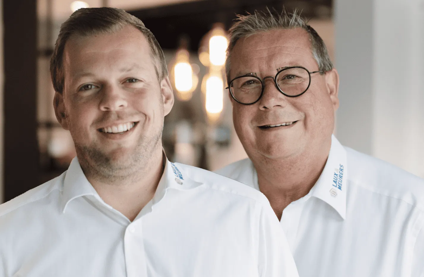 Zwei lächelnde Männer in weißen Hemden mit Firmenlogo, stehend vor unscharfem Hintergrund mit warmem Licht.