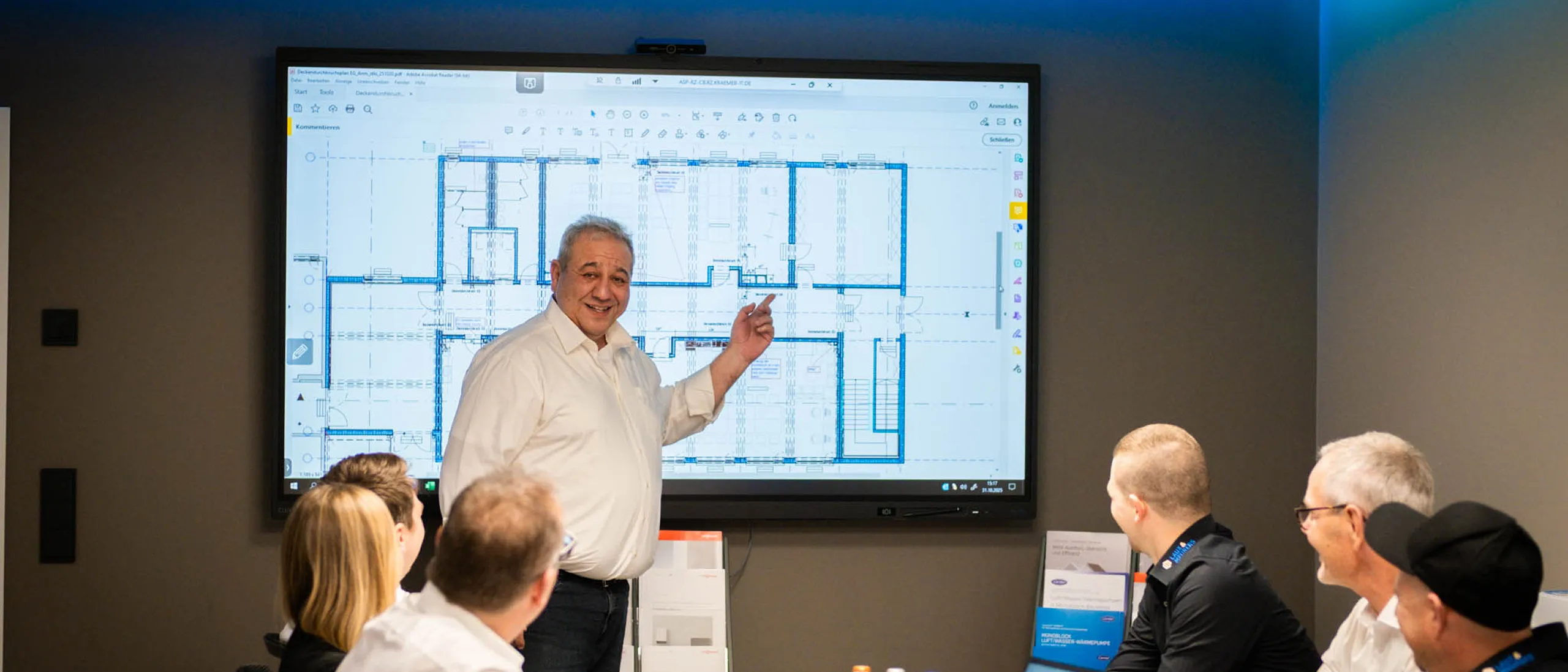 Mann präsentiert Bauplan auf großem Bildschirm vor einem Publikum von fünf Personen in einem Konferenzraum.