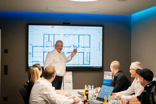 Ein Mann präsentiert einen Architekturplan auf einem Bildschirm vor Kollegen in einem Besprechungsraum.