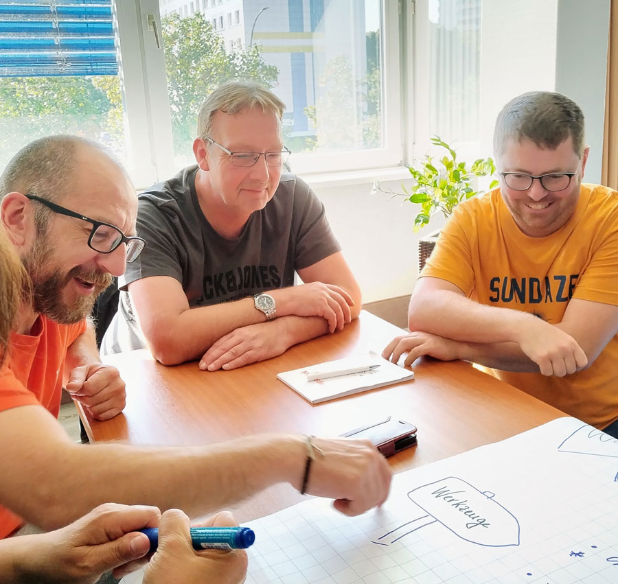 Drei Personen sitzen an einem Tisch und arbeiten gemeinsam an einem großen Blatt Papier.