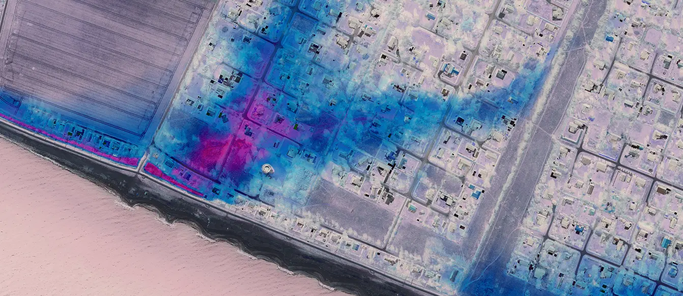 Aerial infrared image of a coastal residential area with visible streets, houses, and agricultural fields near the shoreline.
