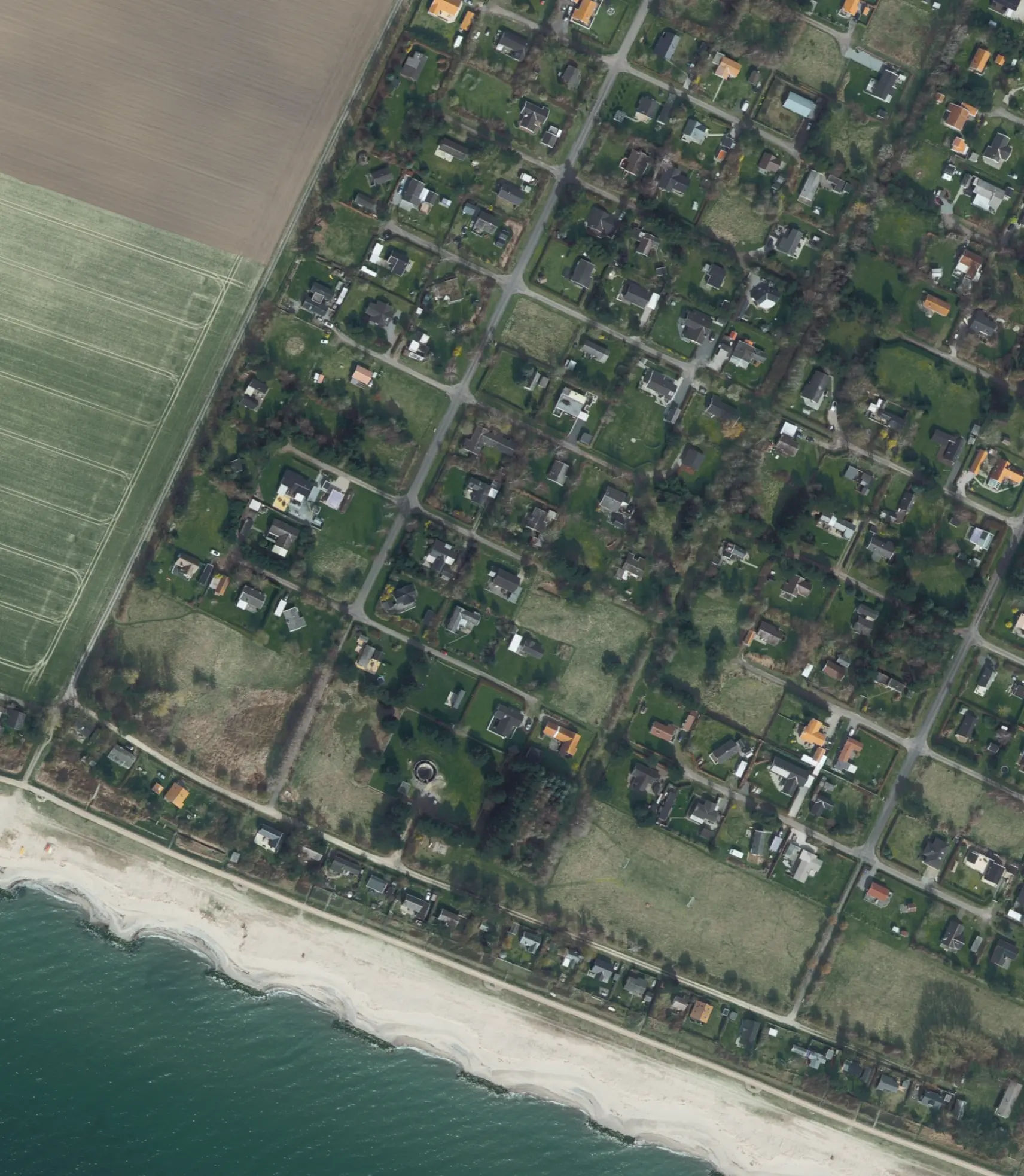 Aerial view of a coastal residential area with a sandy beach, green yards, scattered houses, and adjacent farmland.