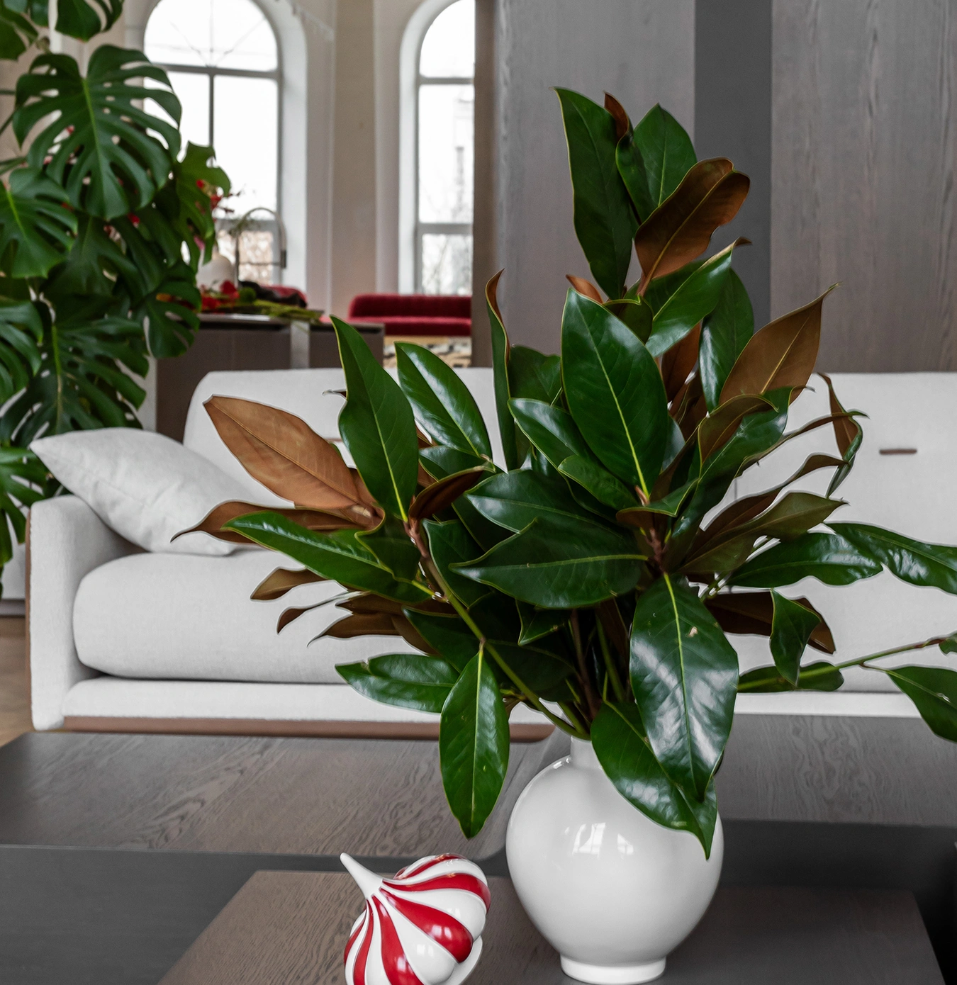A white vase with green and brown magnolia leaves on a wooden table in a modern living room with a white sofa and large windows.