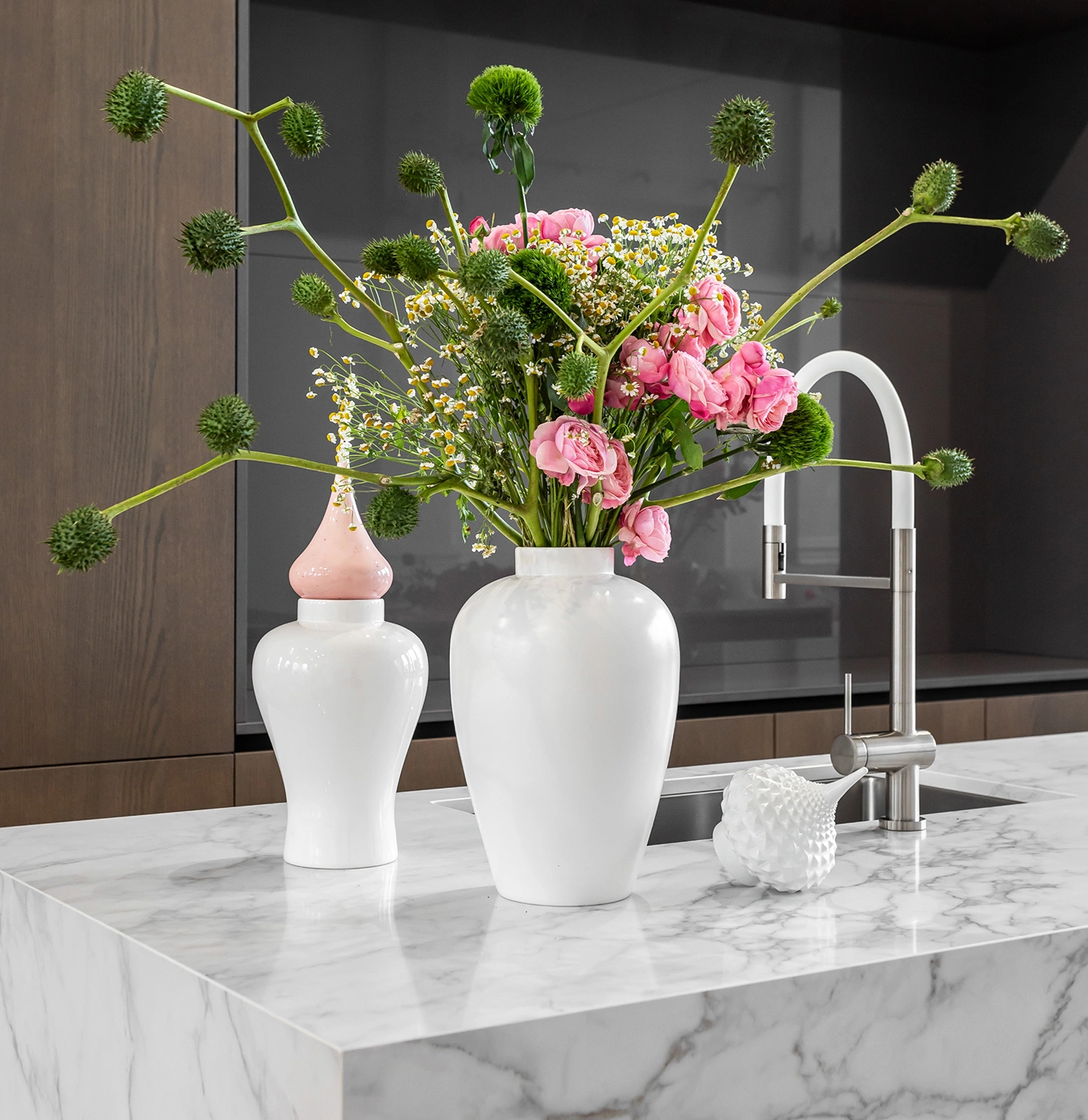 White marble kitchen island with two white ceramic vases, one holding a bouquet of pink roses and green spiky flowers, and a modern stainless steel faucet.