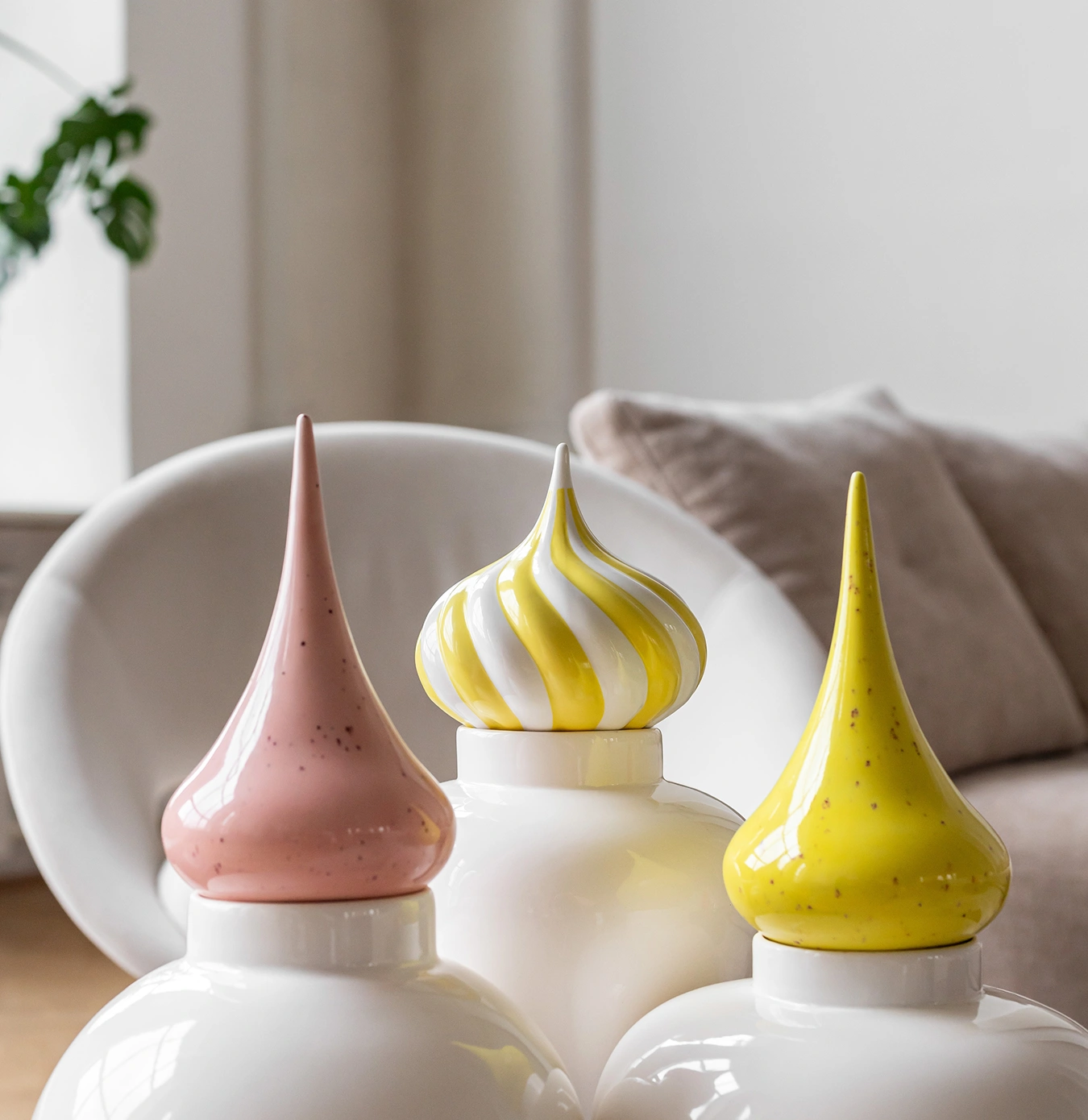 Three white ceramic jars with tall, colorful lids in pink, yellow with speckles, and yellow and white swirl design, arranged on a surface in a cozy living room.