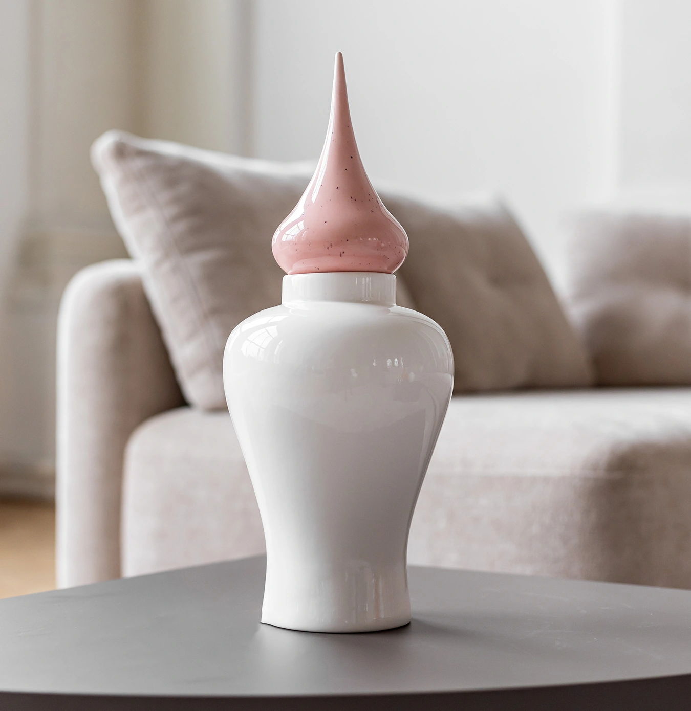 White ceramic decorative jar with a glossy pink pointed lid on a gray table in front of a beige sofa.