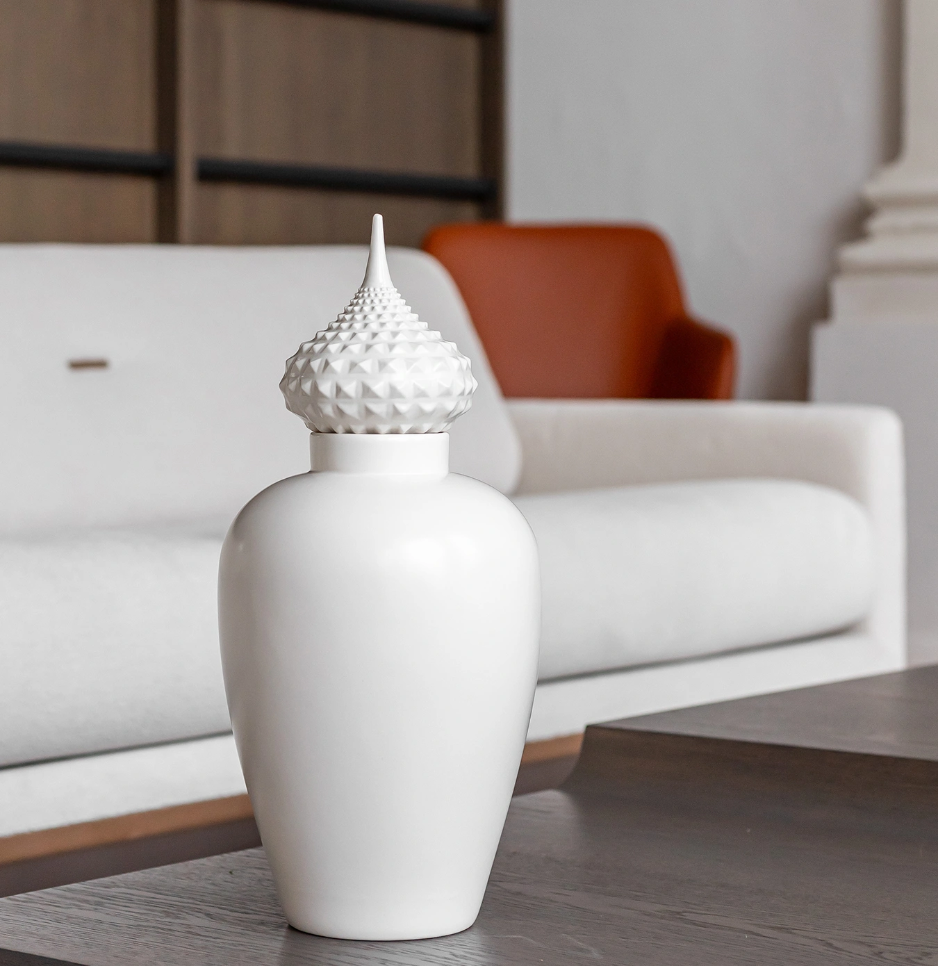 White decorative vase with textured lid on a wooden table in front of a white sofa and brown chair.