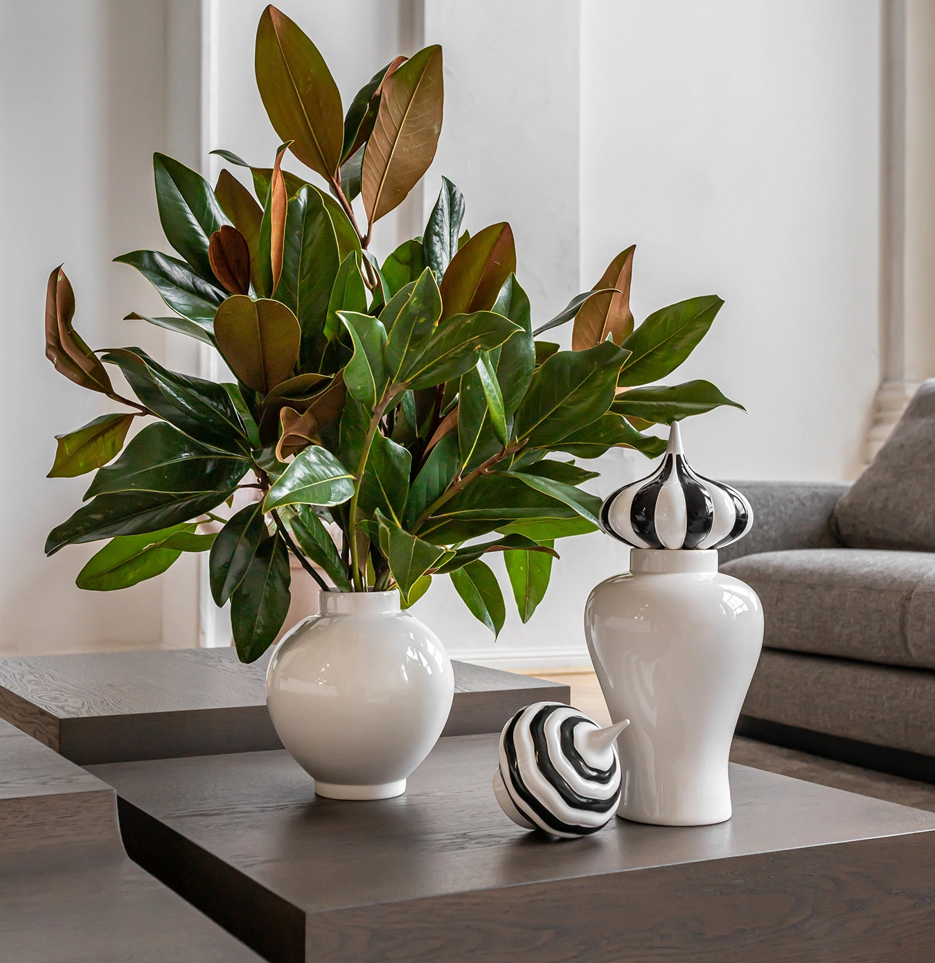 White vase with green and brown magnolia leaves next to a white decorative jar and lid with black and white stripes on a wooden table.