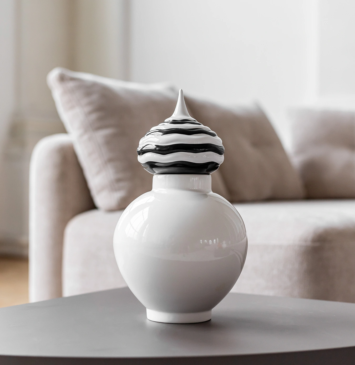 Modern white ceramic vase with a black and white wavy patterned lid on a gray table in front of a beige sofa.