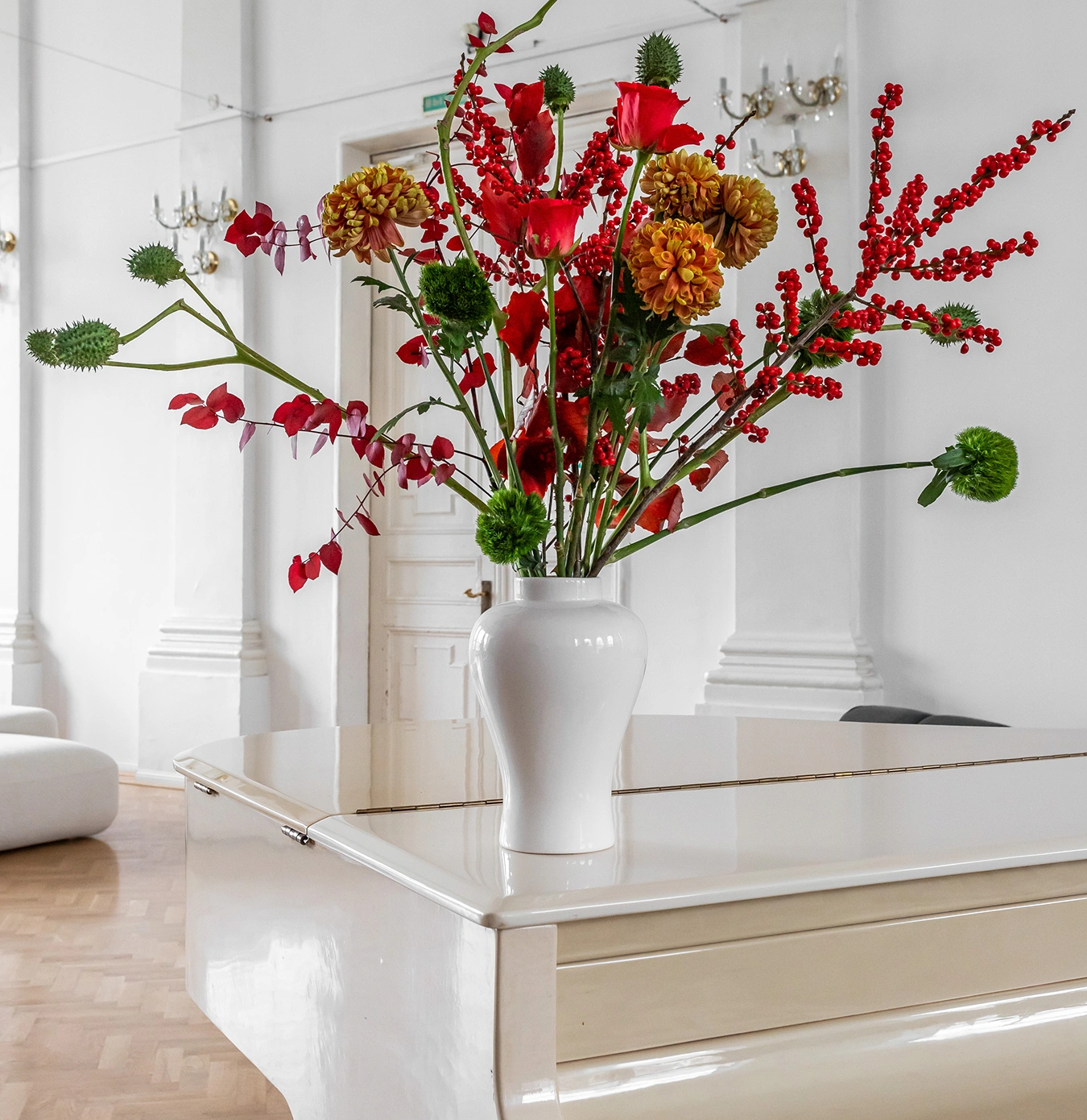 White vase with vibrant red, orange, and green flowers on a glossy white grand piano in an elegant room.
