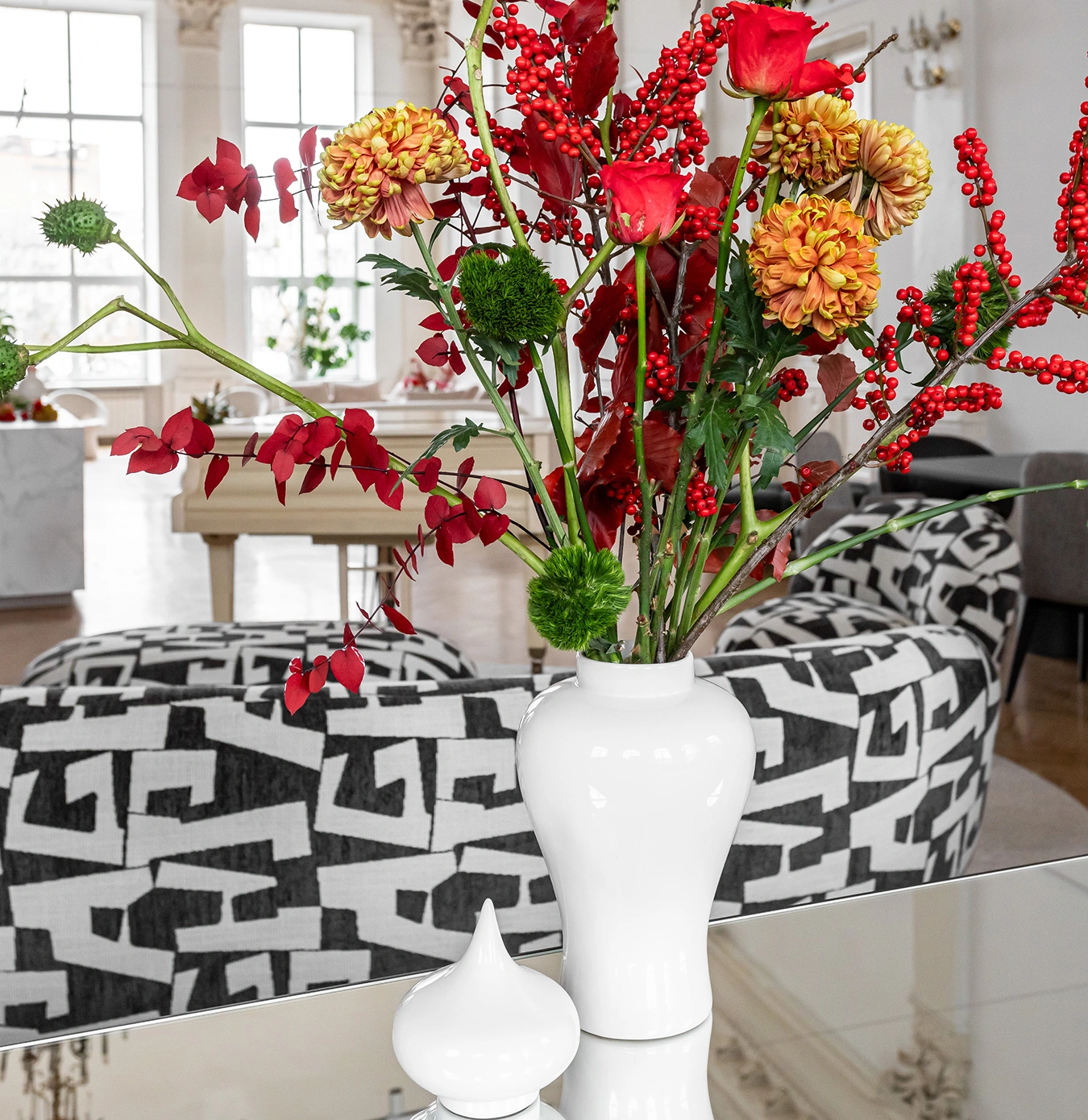White vase with a bouquet of red roses, orange chrysanthemums, green pom-pom flowers, and red berries on a reflective surface in a modern living room.