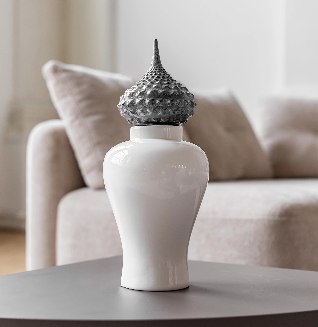 White ceramic vase with a textured metallic pointed lid on a gray table in front of a beige sofa with cushions.