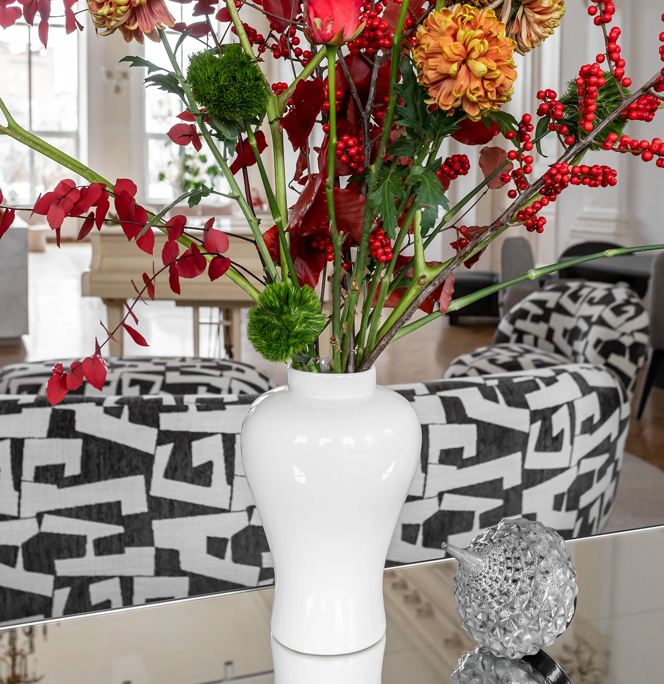 White ceramic vase with red berries, orange chrysanthemums, and green foliage on a reflective surface in front of a black and white patterned sofa.