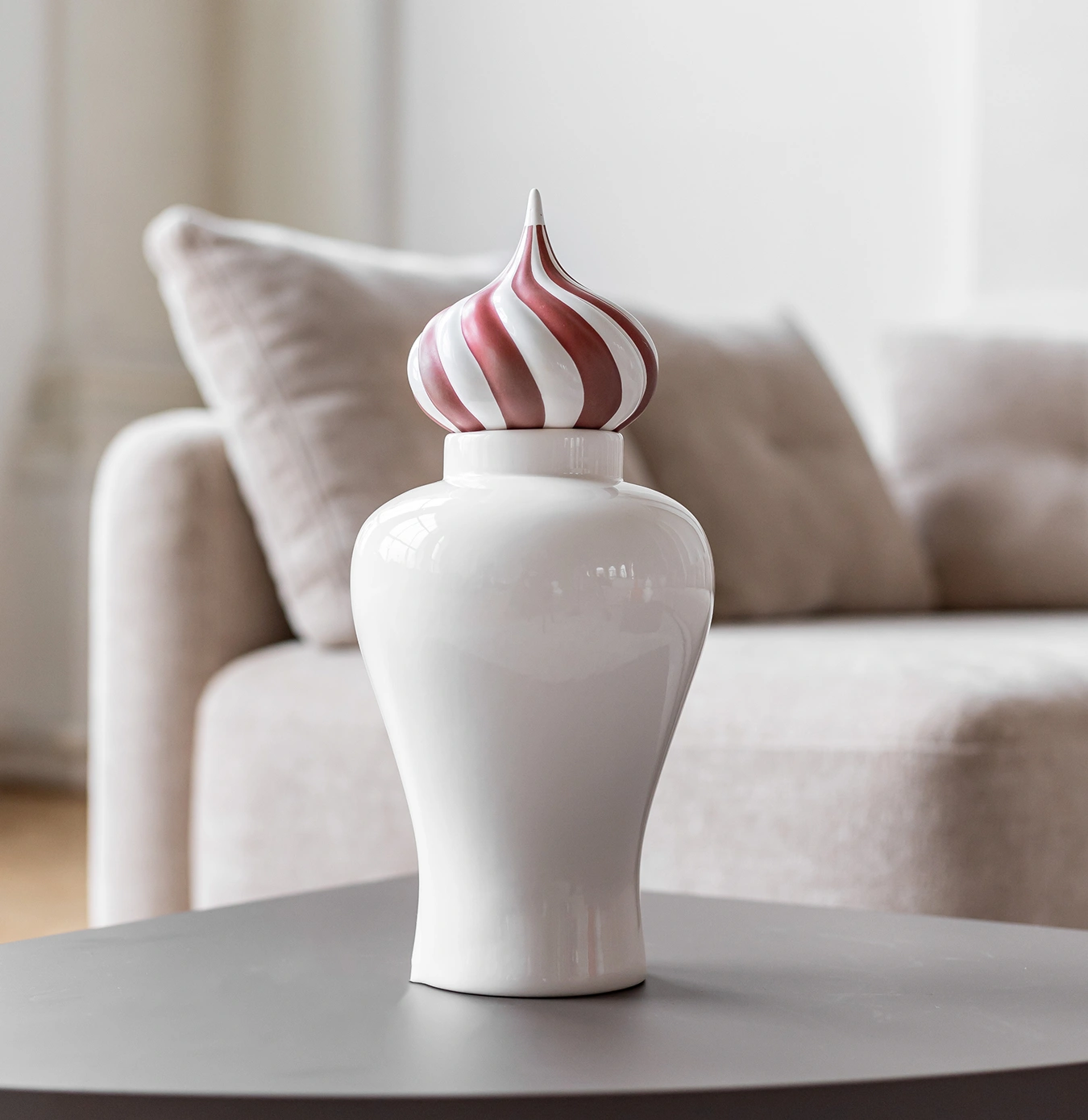 Glossy white ceramic vase with a striped red and white swirl-patterned lid on a black table in front of a beige sofa.