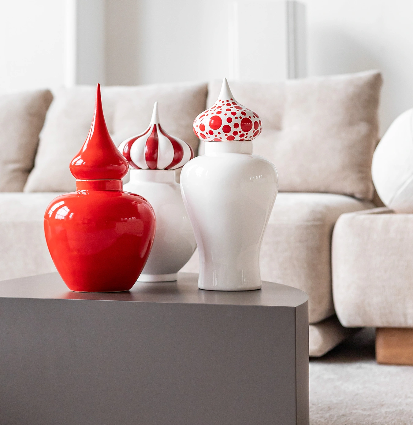 Three decorative ceramic jars with pointed lids in red and white, placed on a gray table in front of a beige sofa.