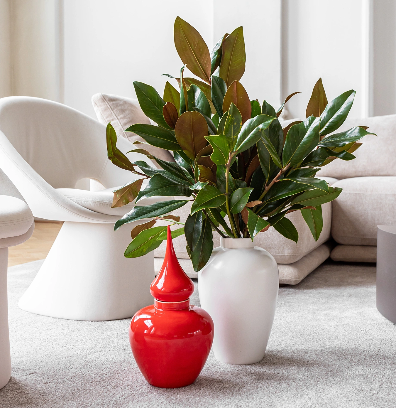 White vase with green leafy branches and a red decorative jar placed on a light carpet in a modern living room.