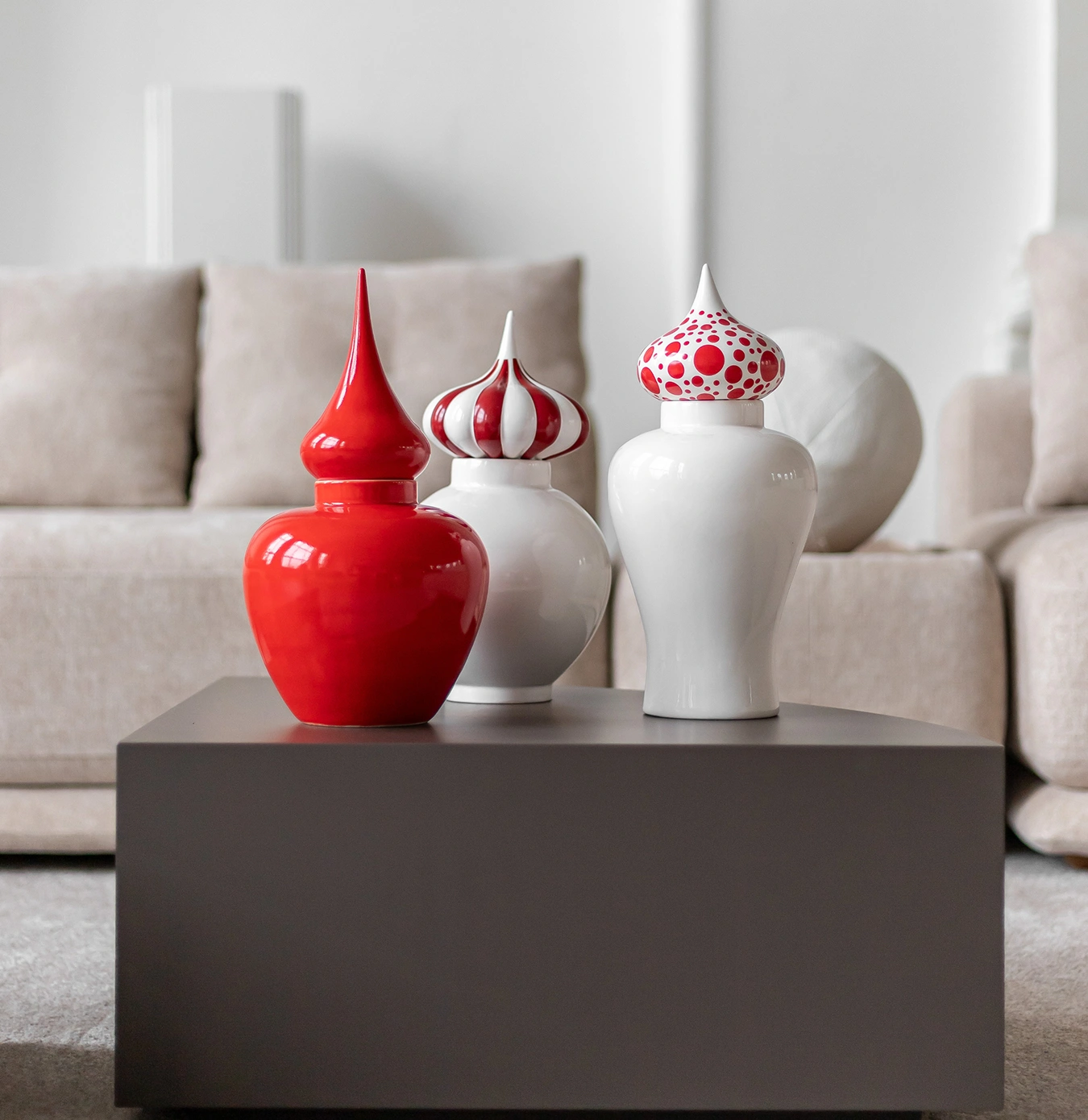 Three decorative ceramic jars with pointed lids, two white with red designs and one solid red, displayed on a dark gray coffee table in a beige living room.