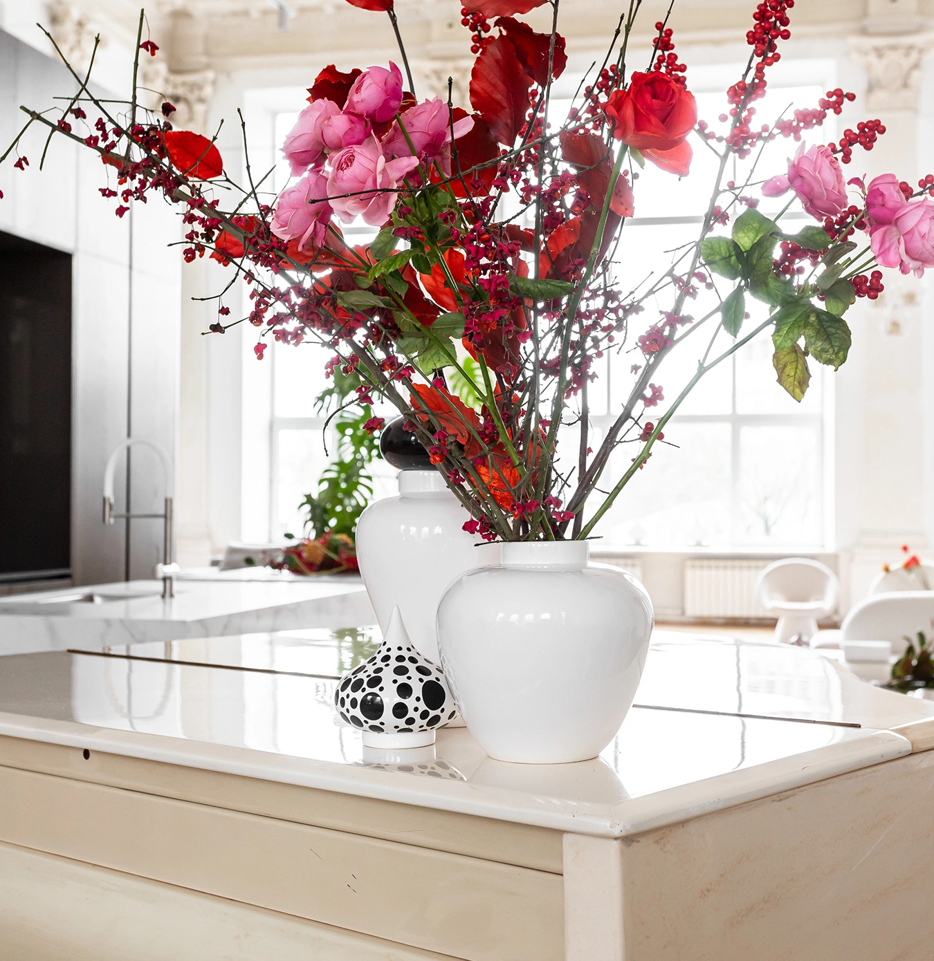 White vases with red and pink flowers on a cream-colored kitchen island with a modern faucet and large windows in the background.
