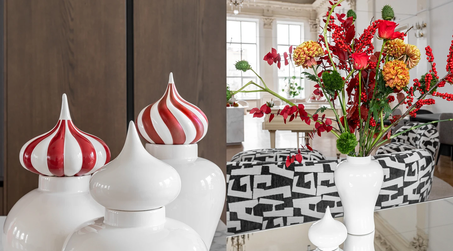 White ceramic jars with pointed lids, two with red and white stripes, displayed next to a white vase holding red and orange flowers and greenery on a reflective surface in a modern living room.