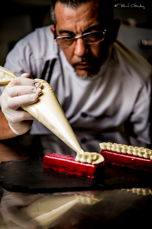 Frédéric Messey en train de décorer un dessert avec une poche à douille, portant des gants. Sur la surface, un gâteau rouge