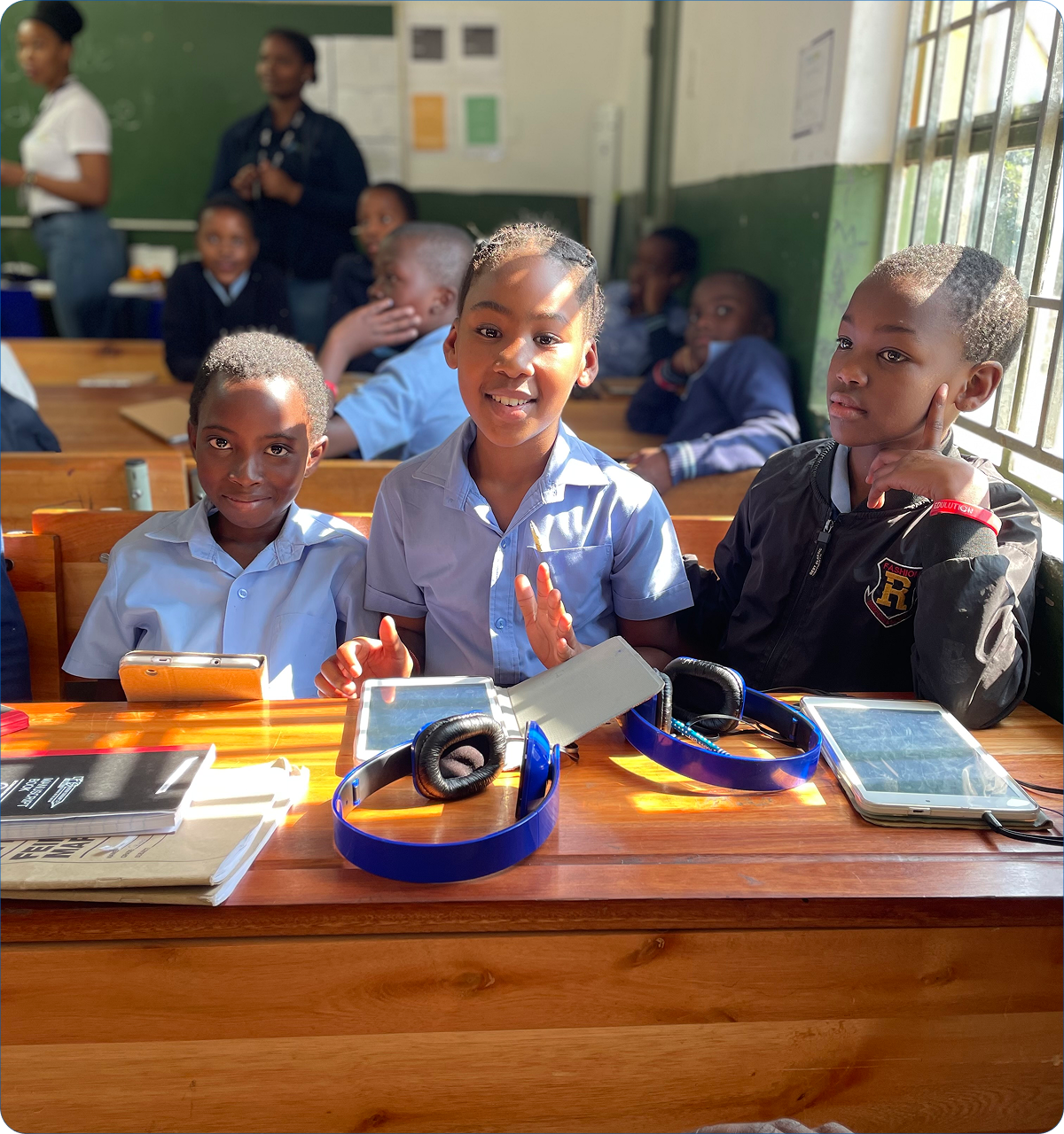 Three children sitting at a wooden desk in a classroom with tablets, headphones, and notebooks, sunlight streaming through a window.