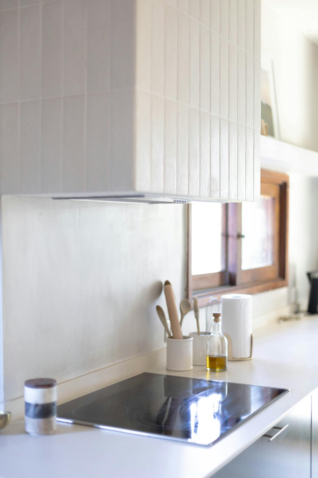 Modern kitchen countertop with electric cooktop, utensil holders, olive oil bottle, and paper towel roll near a window.