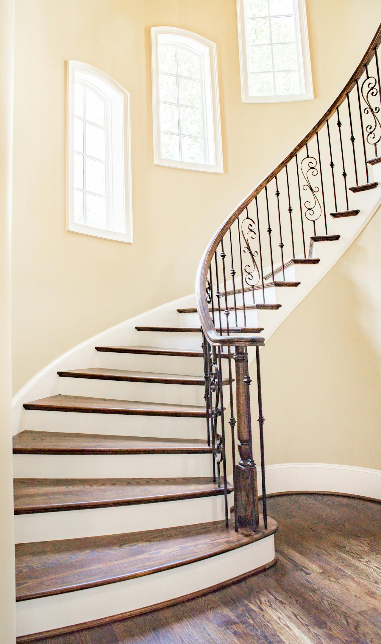 Curved wooden staircase with dark steps and decorative wrought iron railing under three arched windows.
