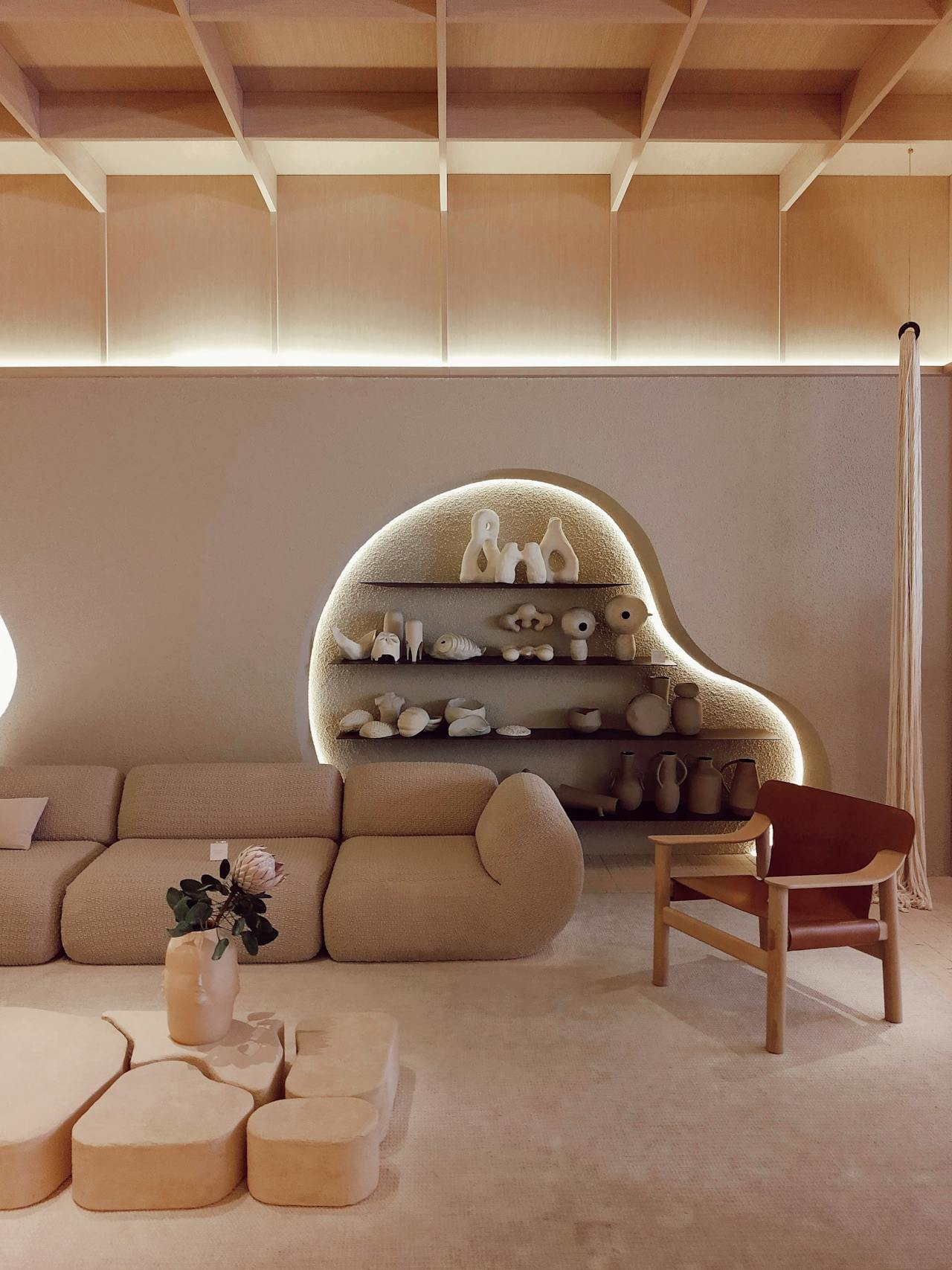 Minimalist living room with beige modular sofa, abstract pebble-shaped coffee tables, wooden chair with brown leather seat, and a curved built-in shelf with ceramic sculptures highlighted by embedded lighting.