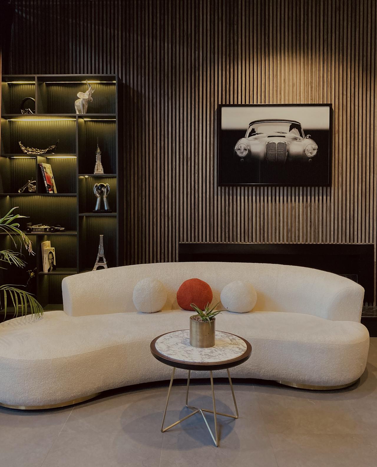 Modern living room with curved white sofa, round marble-top table with a small plant, and a wooden slat wall featuring a black and white vintage car photo and illuminated shelves with decorative items.
