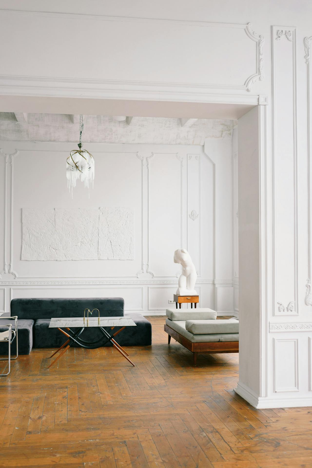 Minimalist living room with ornate white walls, a dark gray sofa, a glass coffee table with wooden legs, a daybed with cushions, and a white abstract sculpture on a small wooden stand.