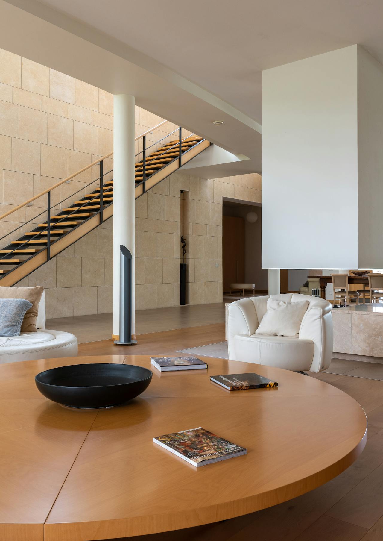 Modern living room with light wood round table holding a black bowl and books, white leather armchairs, and a staircase with black railing.