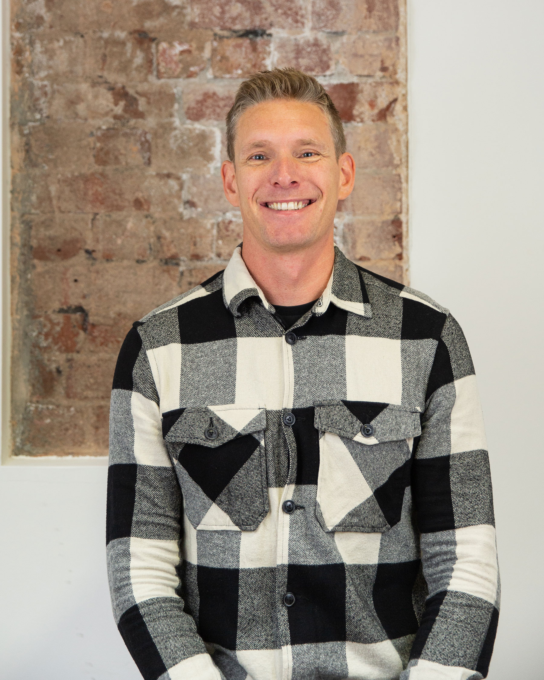 Matt wearing a black and white plaid button-up shirt sitting in front of a brick wall.