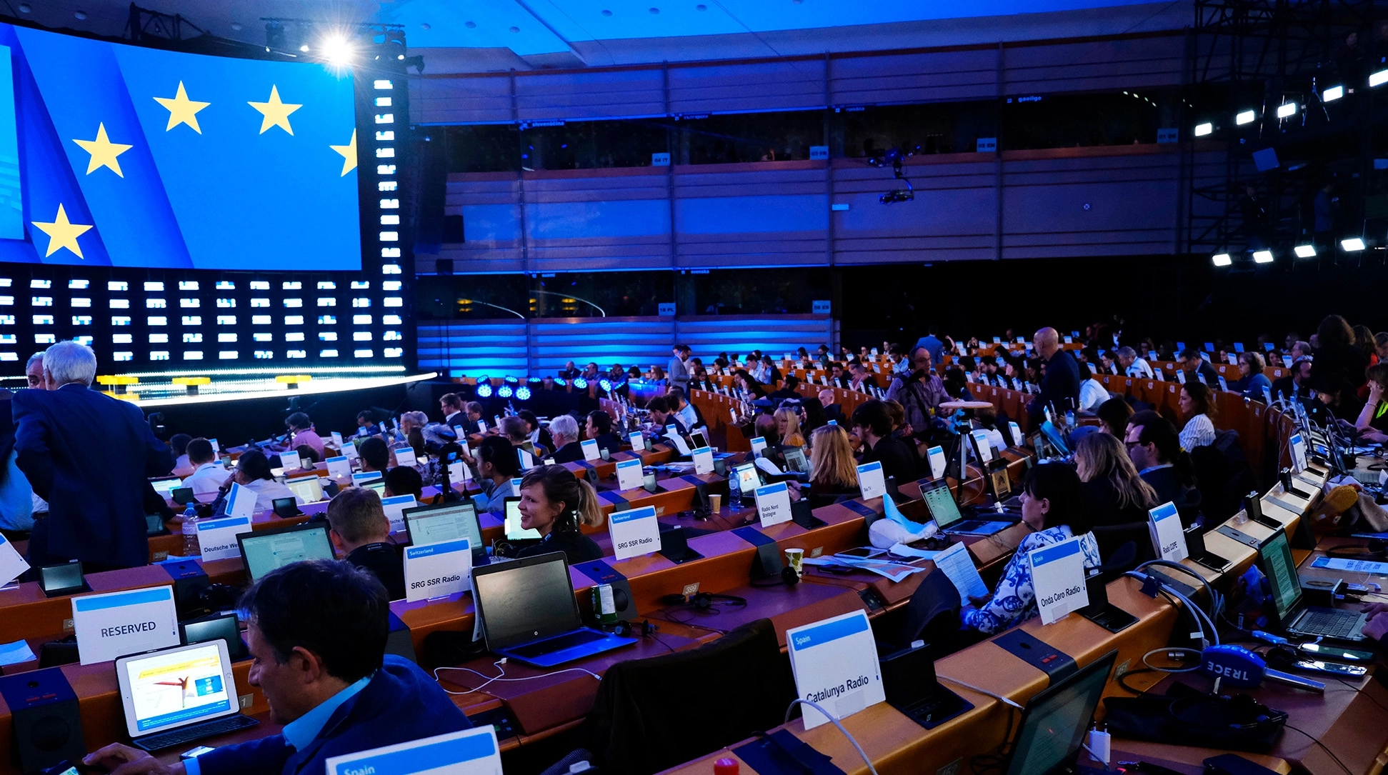 Large conference room filled with delegates seated at desks with laptops and labeled signs, a European Union flag displayed on a screen at the front.