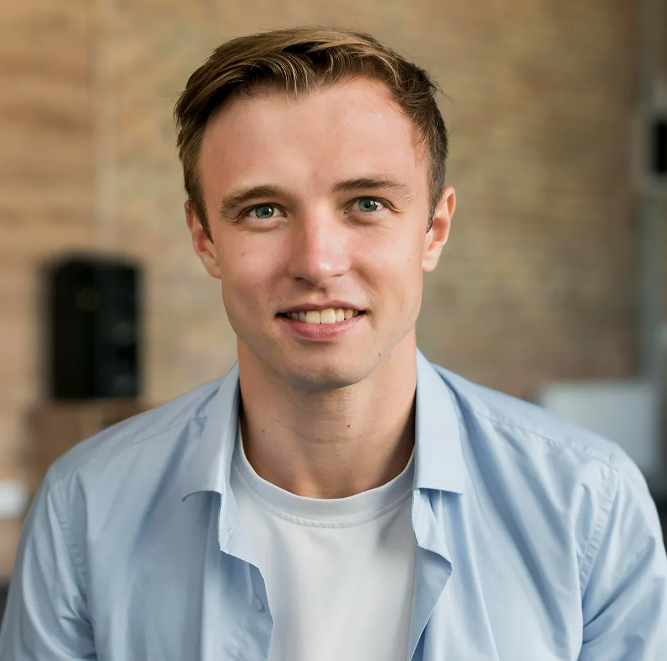Jeune homme souriant portant une chemise bleue claire et un t-shirt blanc avec un mur en arrière-plan flou.