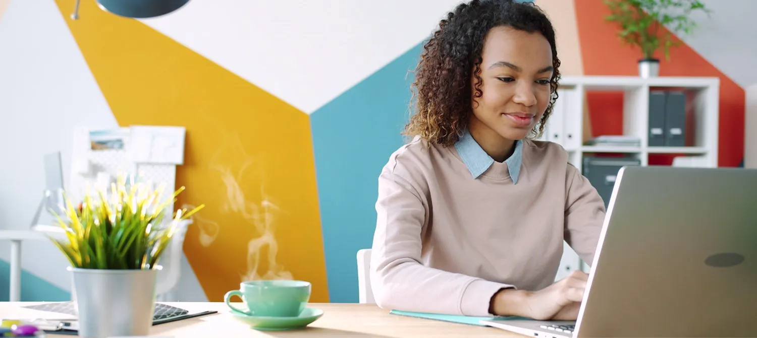 Jeune femme assise à un bureau, travaillant sur un ordinateur portable avec une tasse fumante et une plante en pot sur la table.