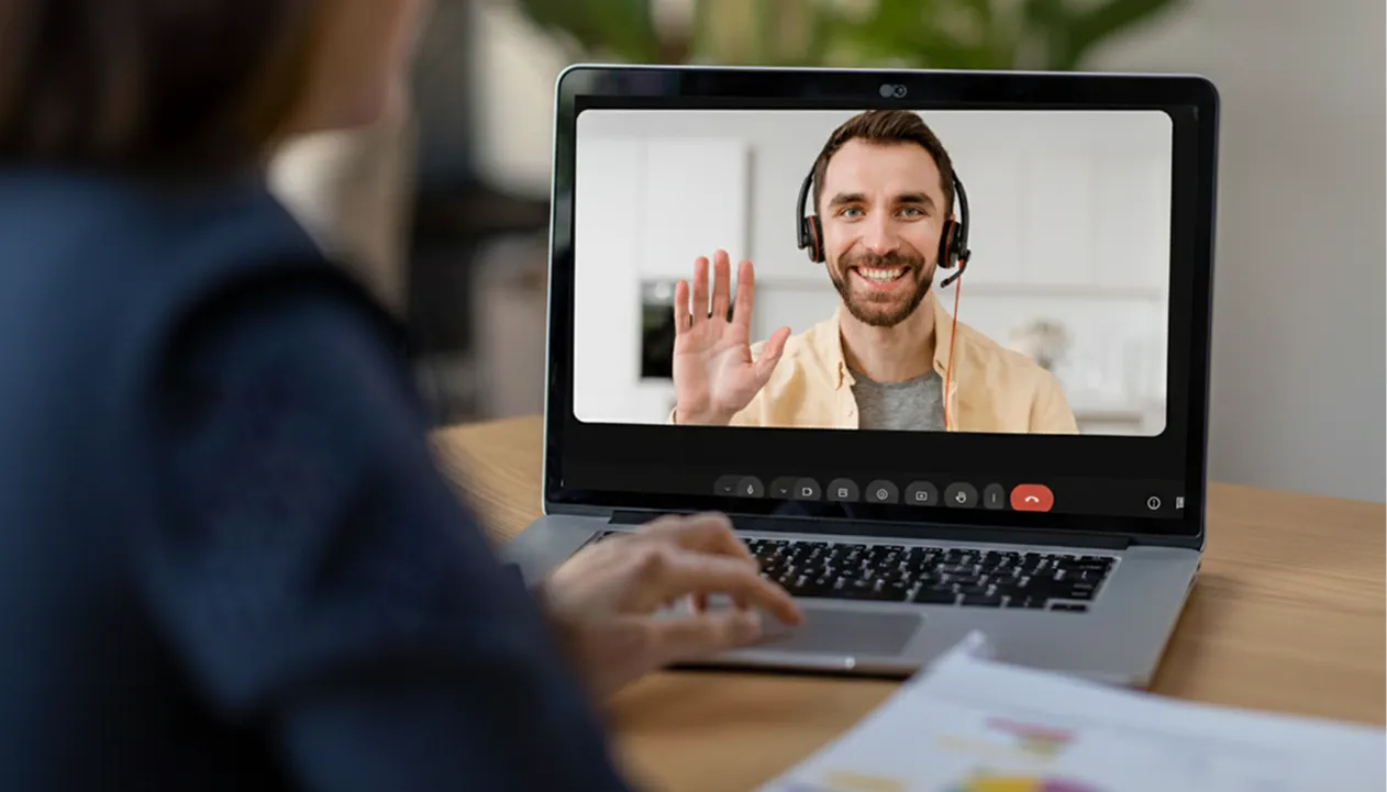 Une femme assise devant un ordinateur portable en visioconférence avec un homme portant un casque qui sourit et lève la main.