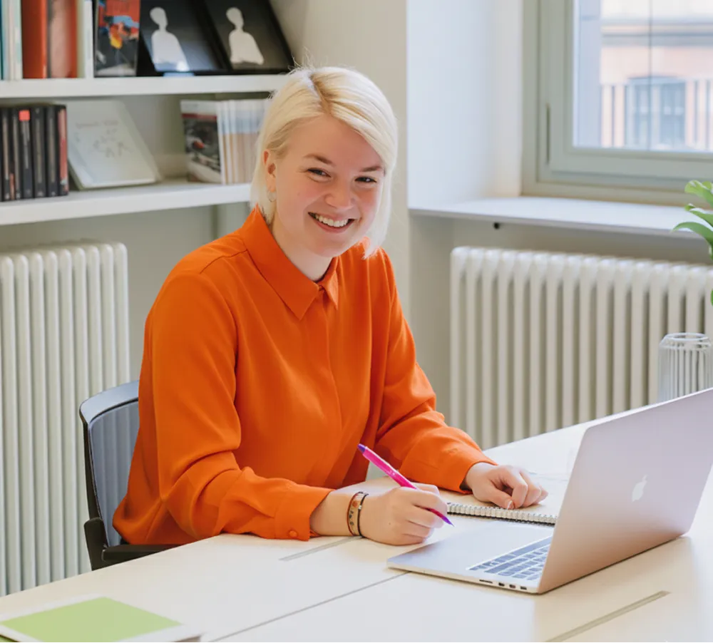 Jeune femme blonde souriante en chemise orange travaillant avec un ordinateur portable dans un bureau lumineux.