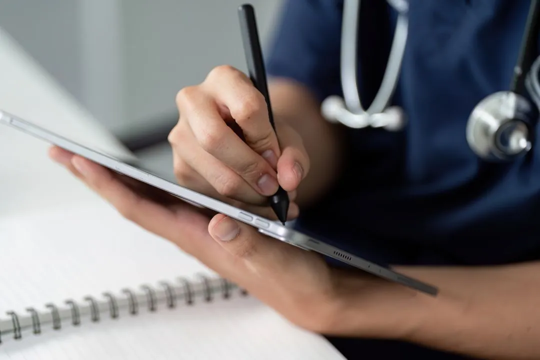 Une personne en tenue médicale utilise un stylet pour écrire sur une tablette numérique avec un carnet à spirale en arrière-plan.