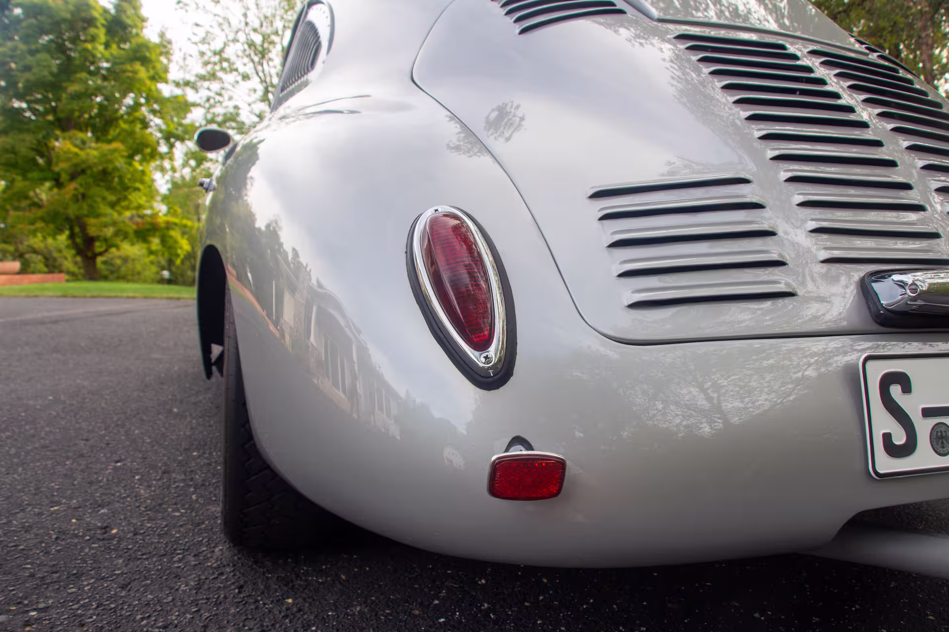 Close-up of the rear end of a silver vintage car showing a red tail light, vented engine cover, and a partial license plate.