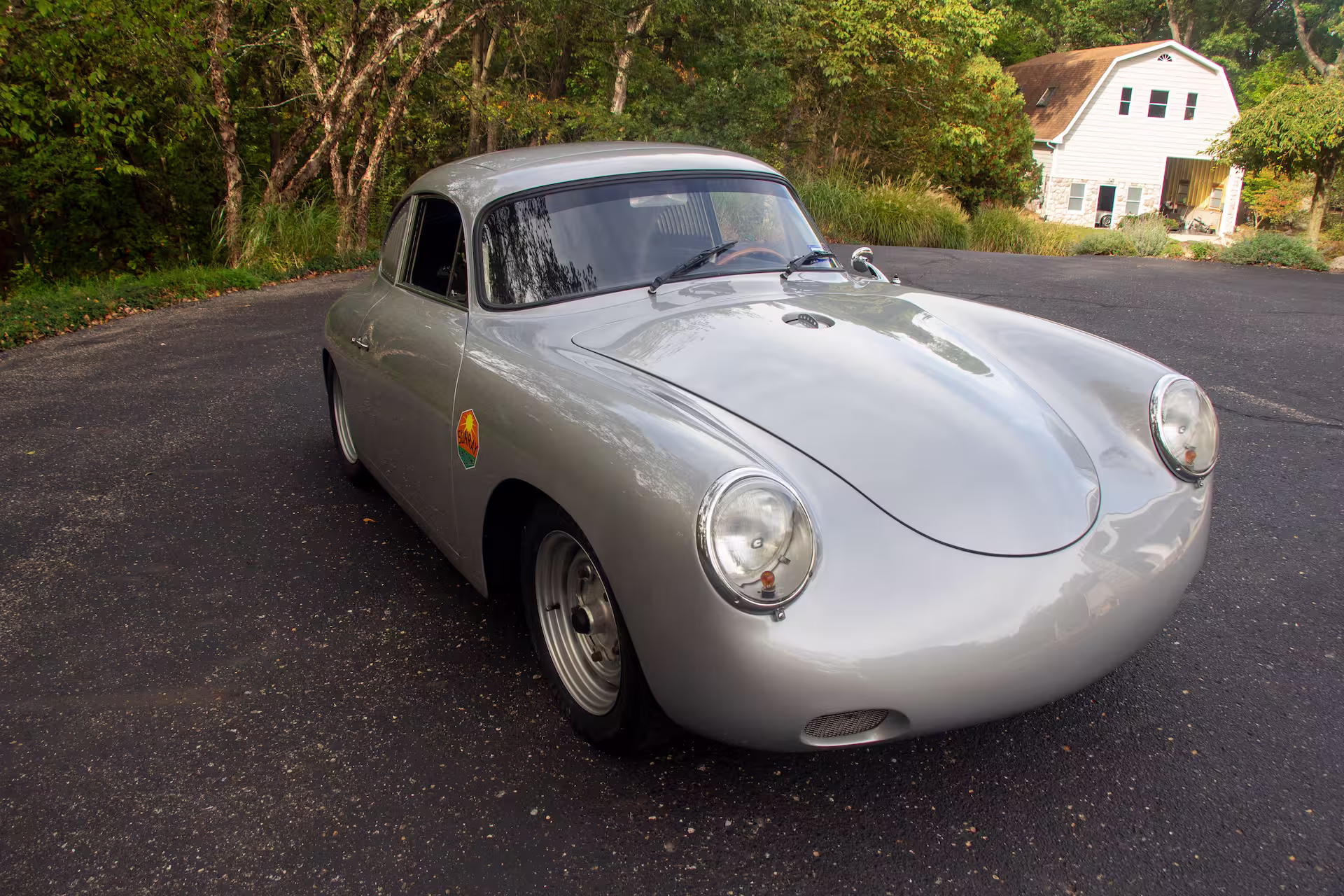 Silver vintage Porsche 356 parked on asphalt driveway near a white house and green trees.
