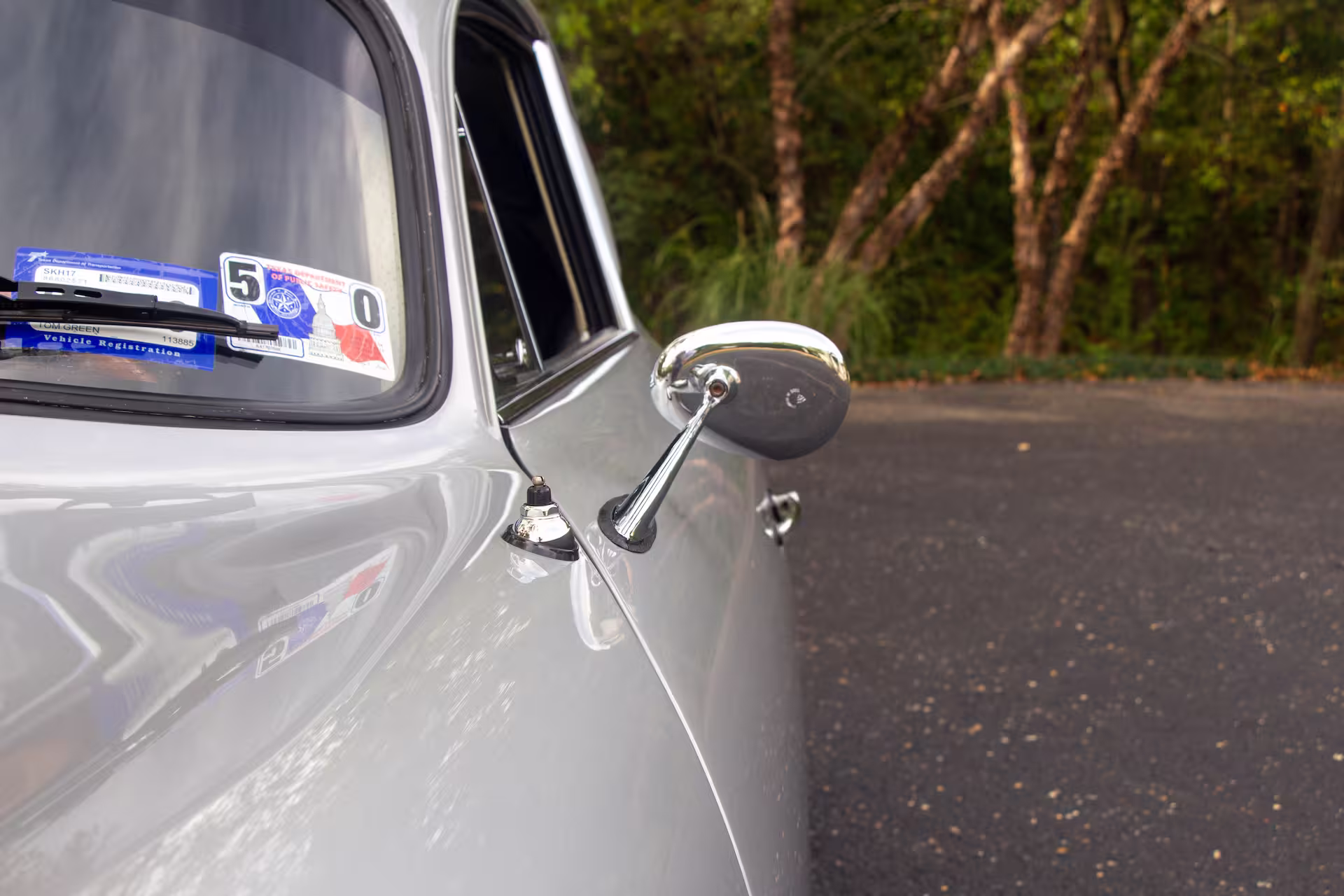 Close-up of a silver vintage car's side rearview mirror and partially visible windshield with registration stickers.