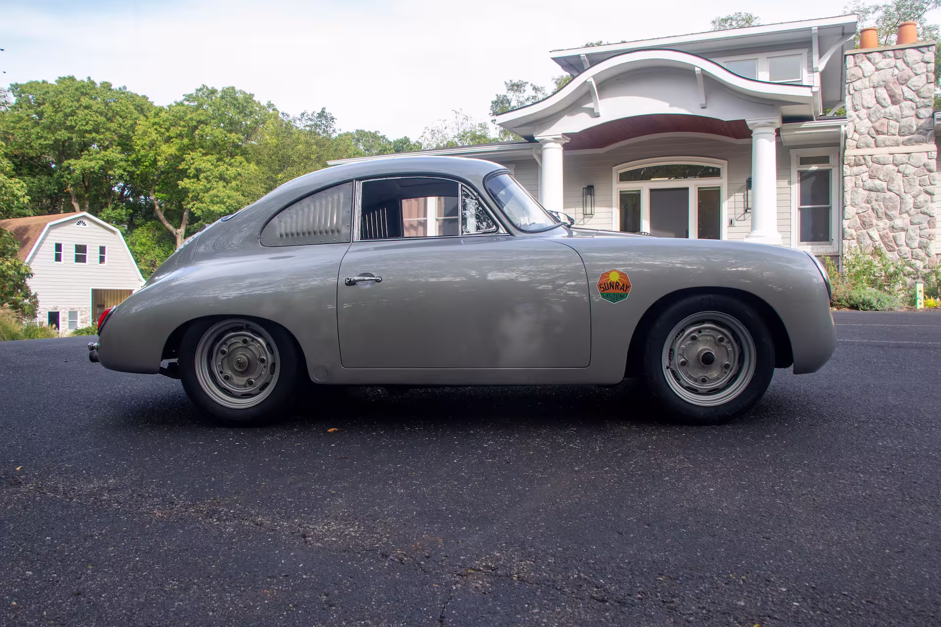 Side view of a classic silver Porsche 356 parked on a driveway in front of a white house with columns.