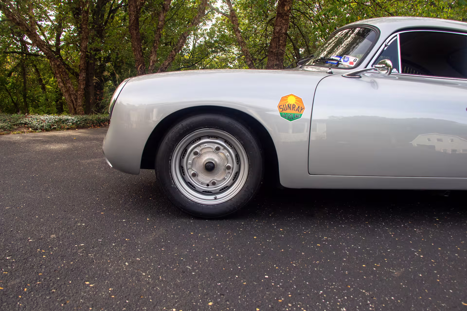 Side view of a silver classic car showing the front wheel and a 'Sunray Gasoline' sticker on the door with trees in the background.