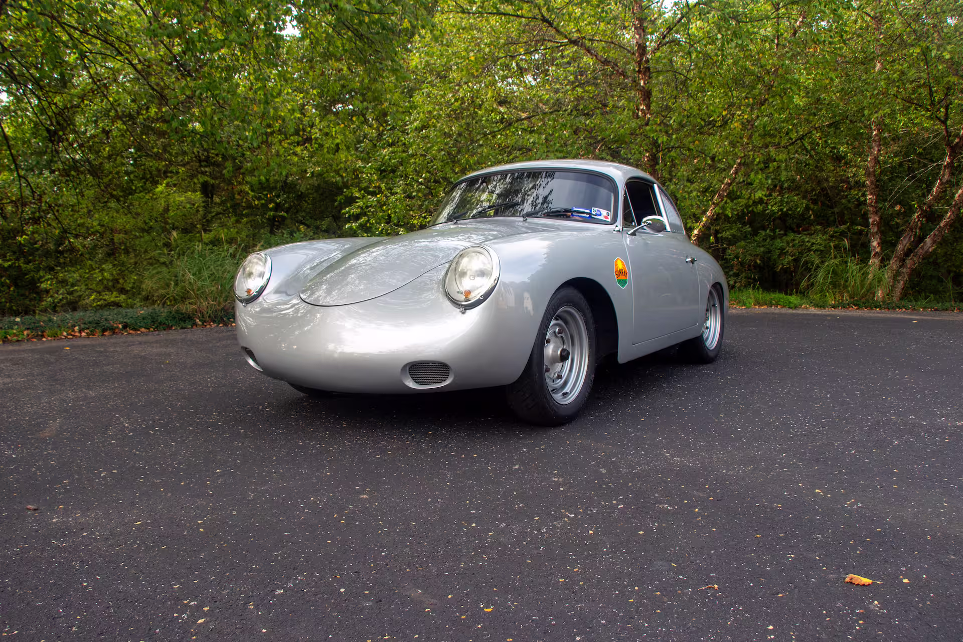 Silver vintage Porsche 356 car parked on an asphalt road with green trees in the background.