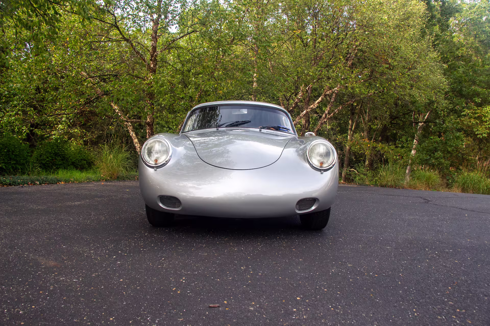 Front view of a silver vintage sports car parked on asphalt with green trees in the background.