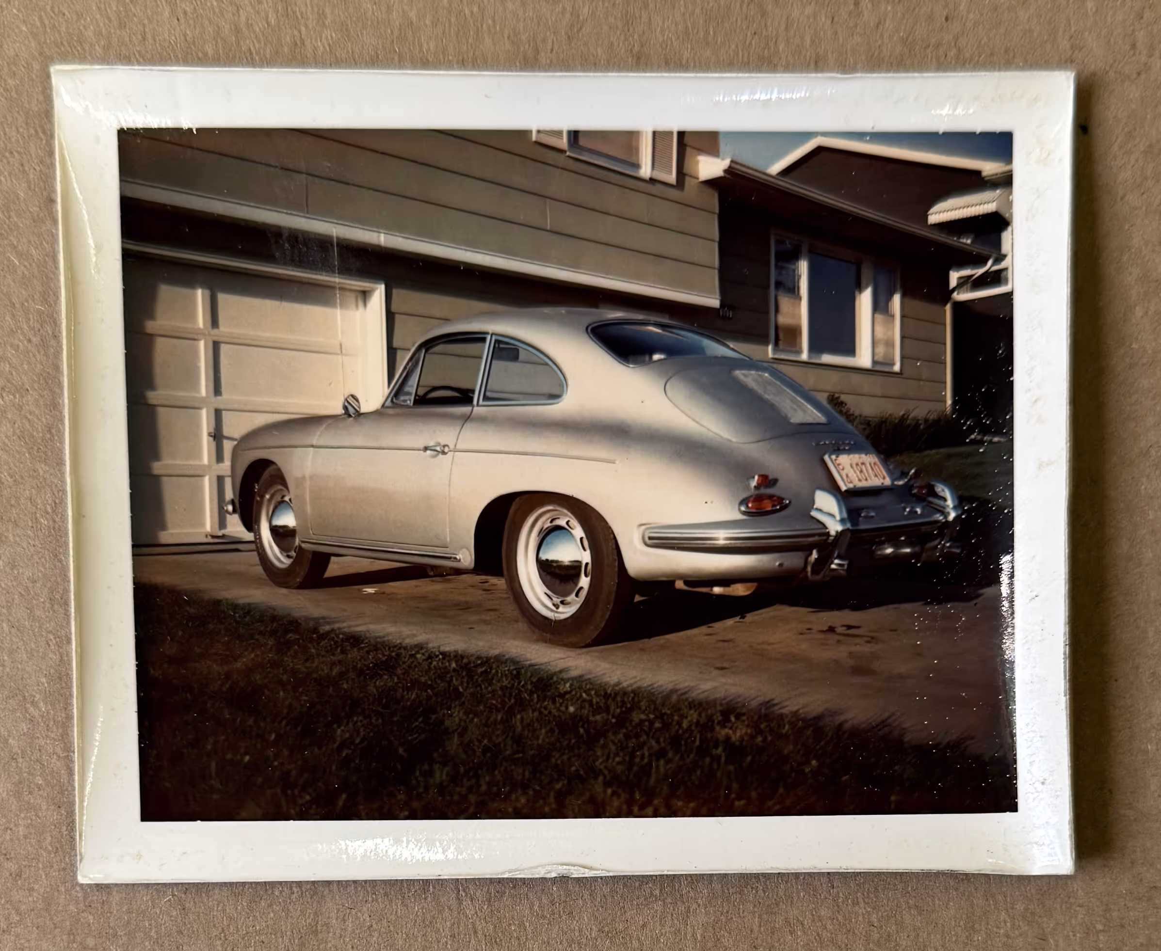 Vintage silver Porsche 356 parked on a driveway in front of a house garage.