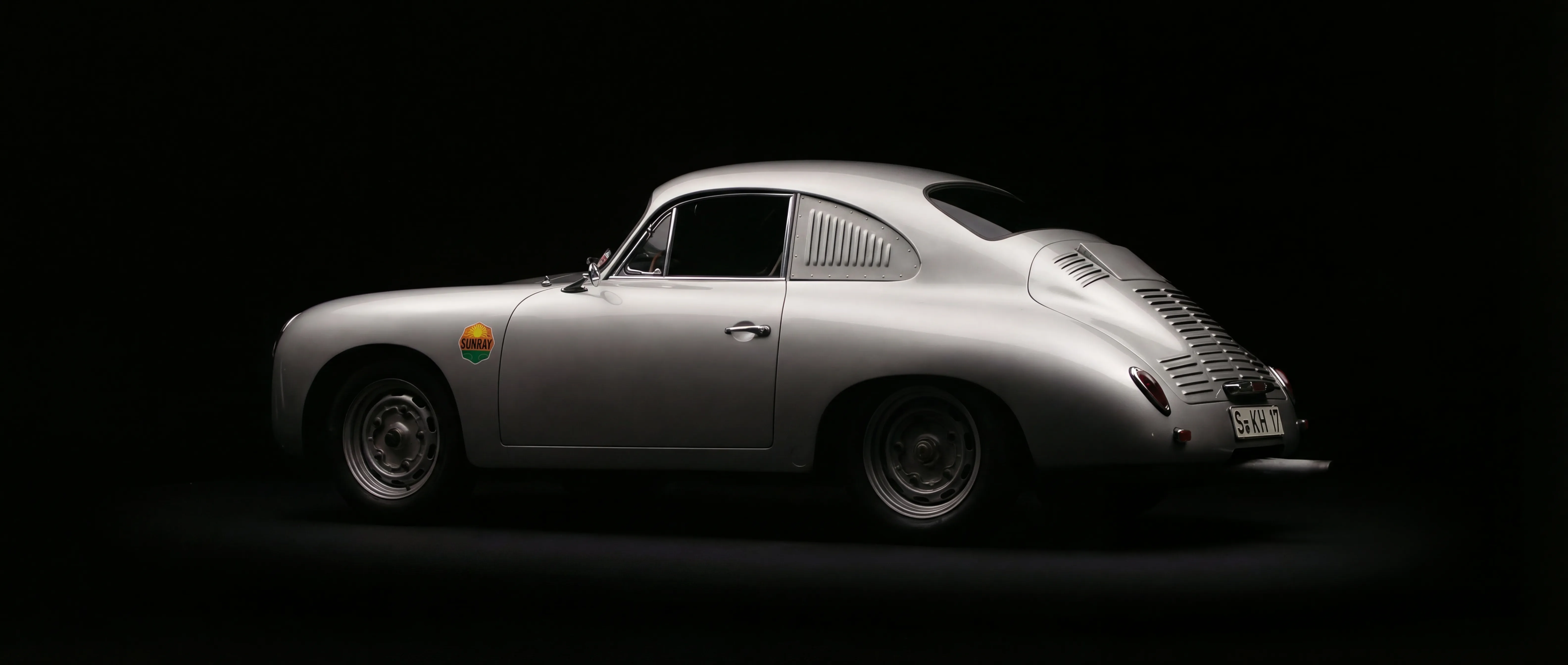 Side view of a classic silver Porsche 356 with vintage Sunray decal on front fender against black background.