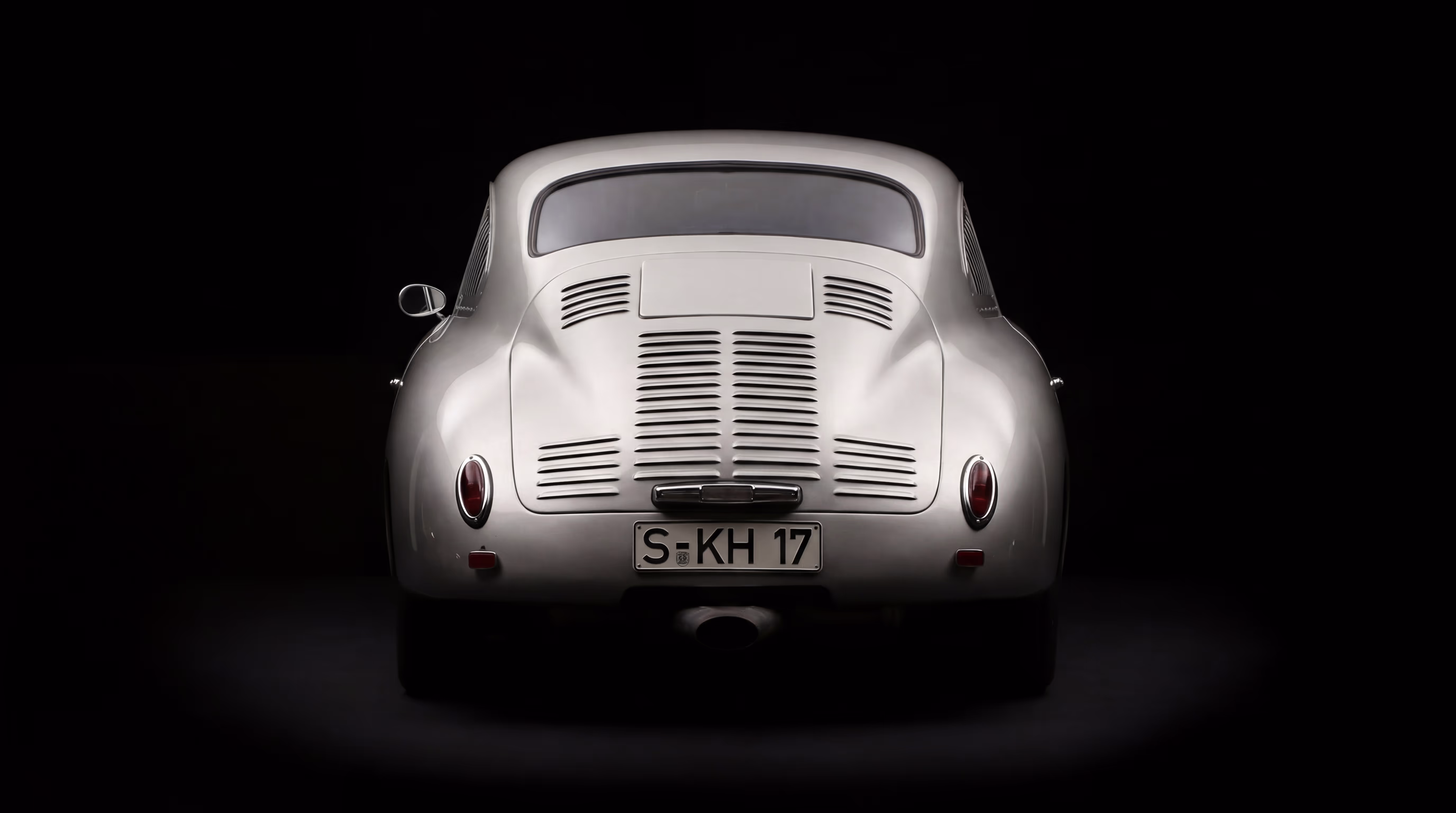 Rear view of a classic silver Porsche car with vintage tail lights and ventilation grilles on a black background.