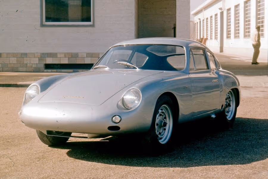 Silver vintage sports car parked on a street with a building and a person in the background.